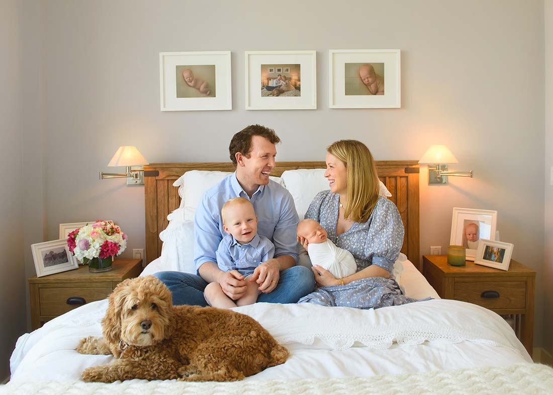 A smiling family of four sits on a bed, captured by a Manhattan family professional photographer. The mother holds a newborn, the toddler smiles beside them, and their brown dog relaxes at the foot of the bed amid cherished family photos.