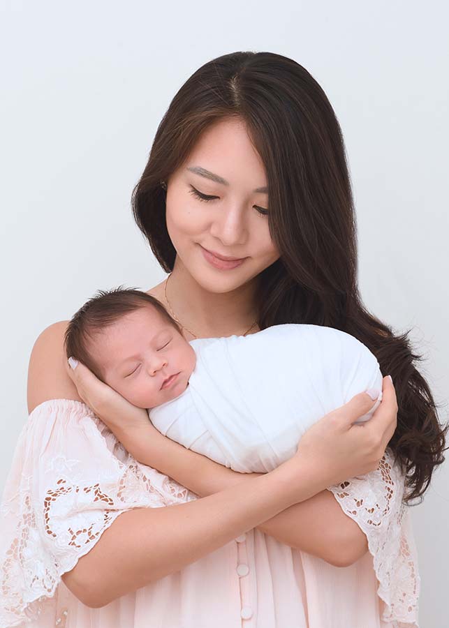 A mother gently cradles her baby boy, both in soft, light-colored clothing, against a plain light background—a tender infant portrait capturing Upper East Side elegance.