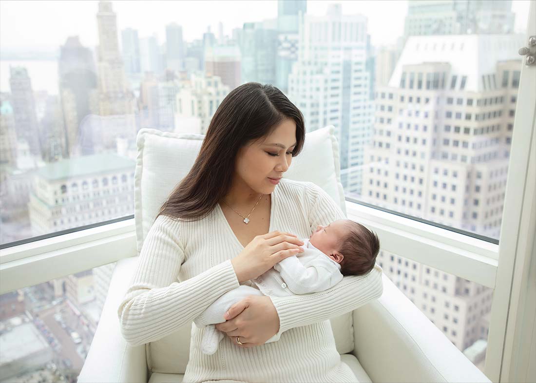 A woman in a white sweater sits in a bright room by large windows, cradling her newborn baby. The Manhattan view in-home newborn photoshoot captures the city skyline with tall buildings just beyond the glass.
