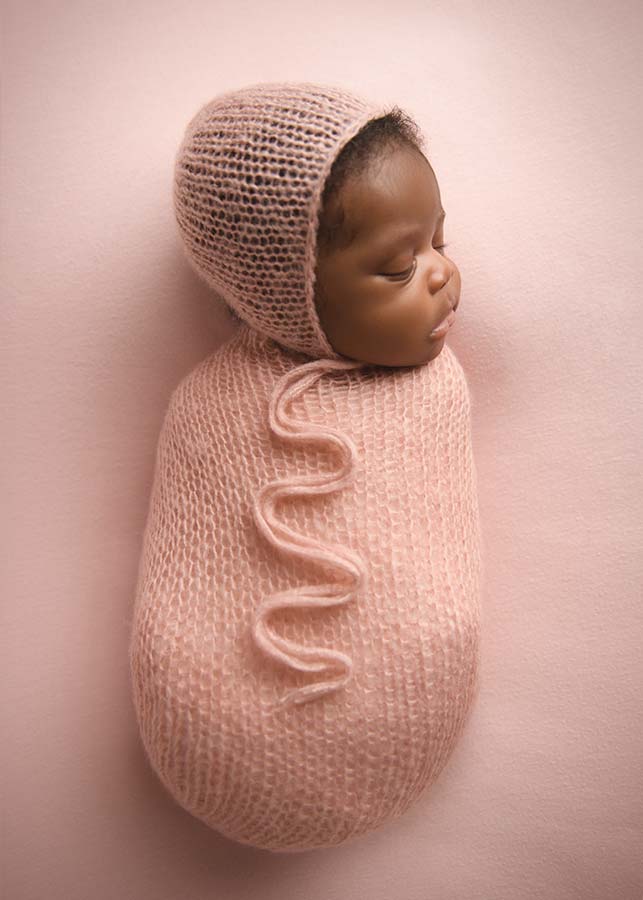 Newborn baby peacefully sleeping, swaddled in a soft, knitted pink blanket with a matching bonnet, on a pale pink background.