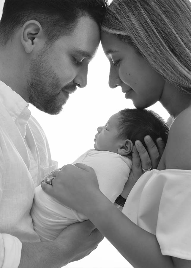 A man and a woman gently touch foreheads while holding a sleeping newborn baby wrapped in a blanket, all smiling softly in a tender, loving moment during an in your home baby photoshoot in New York. The image is in black and white.