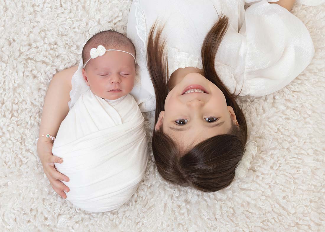 A young girl lies on a soft white rug, smiling up at the camera, with her arm around a swaddled newborn baby in a headband. This heartfelt siblings photo session in Battery Park City captures their sweet bond as both children are dressed in white.