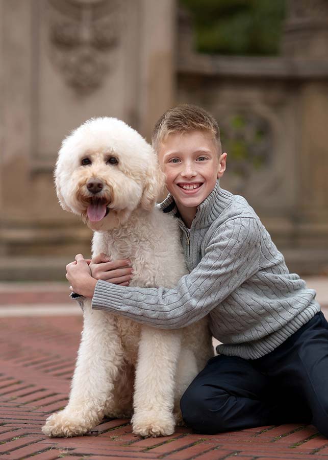 A smiling boy kneels on red bricks at Bethesda Terrace in NYC, hugging his fluffy, light-colored pet dog. The cheerful pup sits and pants, while a stone wall creates a softly blurred background.