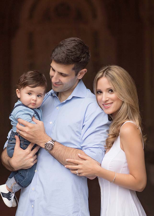 A smiling mother in a white dress stands beside the father in a light blue shirt, holding their son. The family poses closely together in Central Park, with the baby looking at the camera and the background softly blurred.