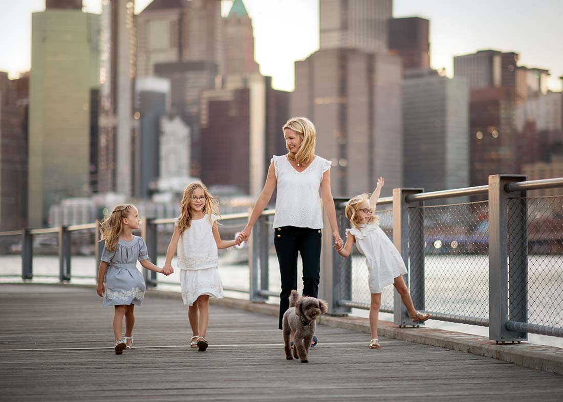 A mother walks on a boardwalk near the Brooklyn Bridge, holding hands with her daughters and walking their small gray pet dog. The city skyline rises in the background as the group appears happy and relaxed.