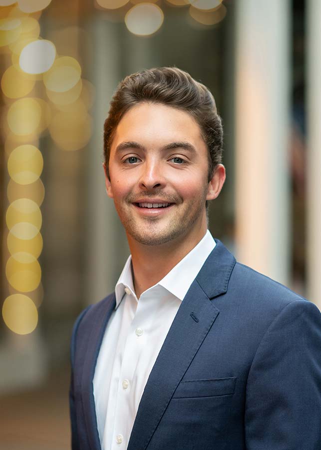 A college senior with short brown hair, wearing a navy blue suit and white shirt, smiles at the camera. The softly blurred background features warm, circular lights, suggesting an elegant evening at the Plaza Hotel NYC.