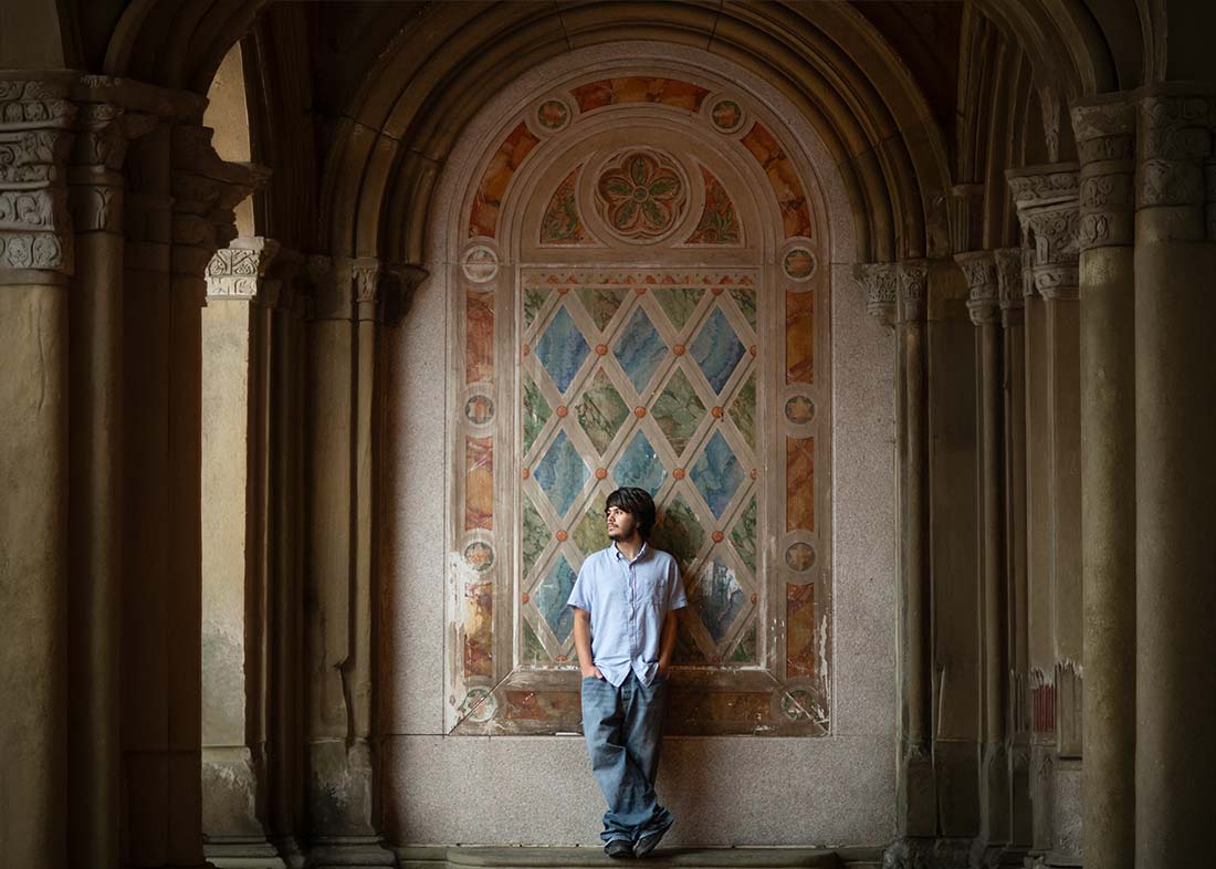 A man in a light blue shirt and jeans stands casually against an ornate, arched wall with a colorful, patterned window in the dimly lit Bethesda Terrace alcove—perfect for a striking senior photoshoot look as he gazes upward to his right.