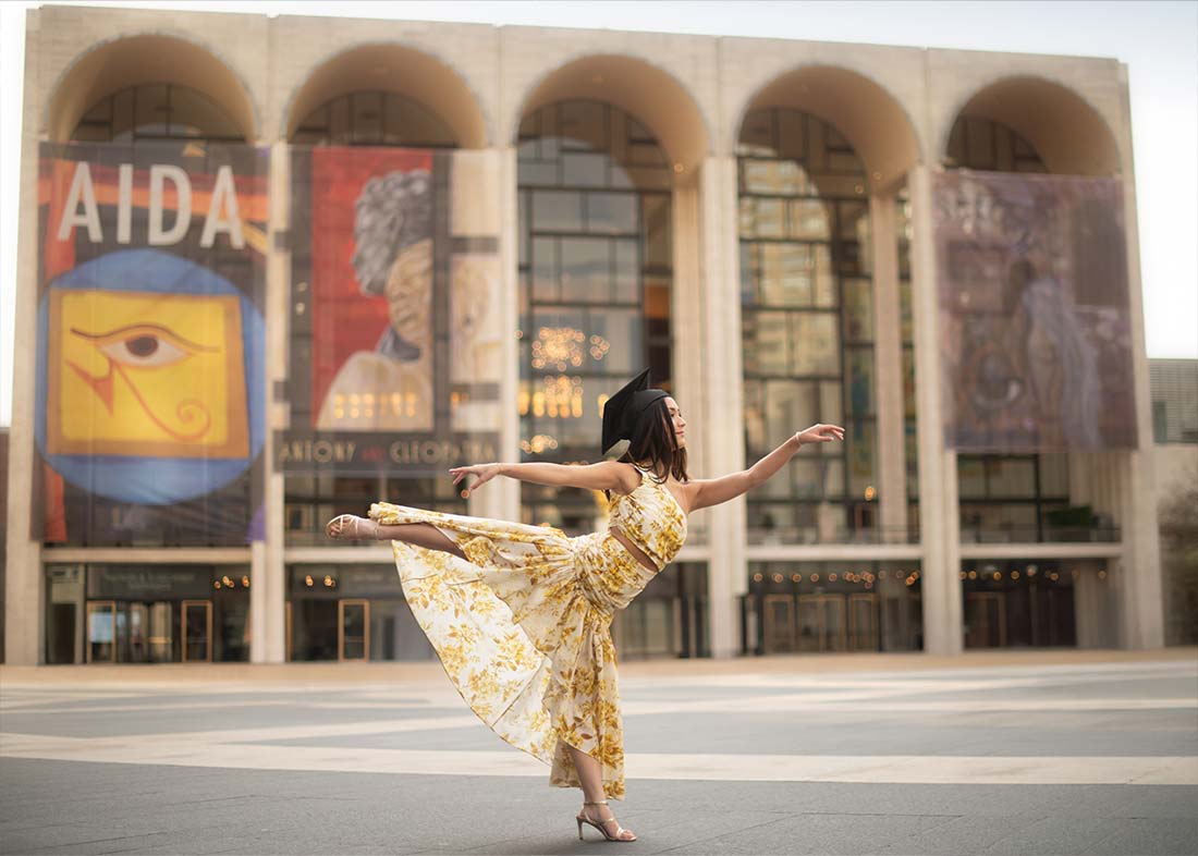 A Juilliard School graduate in a yellow floral dress and graduation cap gracefully poses in a ballet stance outside Lincoln Square's grand theater, framed by tall arches and colorful banners.