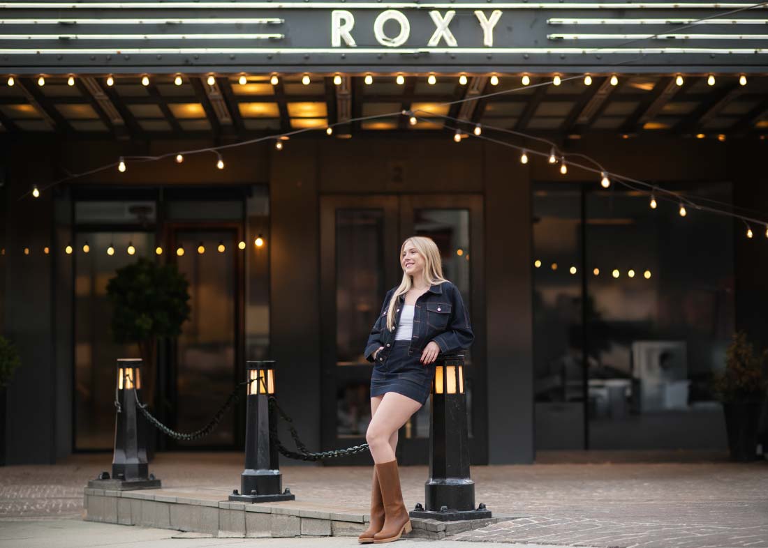 A woman in a dark jacket, skirt, and brown boots stands outside under string lights in front of the Roxy Hotel NYC, the perfect spot for a stylish senior portrait.