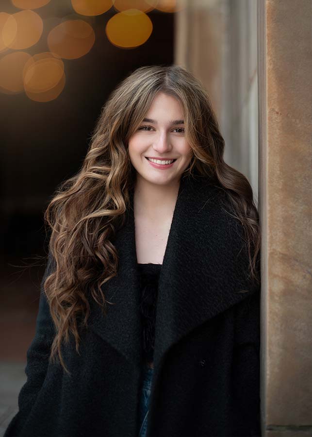 A high school senior with long wavy brown hair, wearing a black coat, smiles while leaning against a wall. The background is softly blurred with warm bokeh lights.