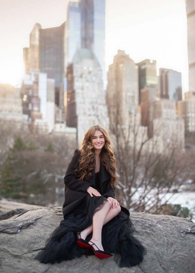 A young woman with long, wavy hair sits on a rock in a black dress and red shoes, smiling during her Manhattan skyline senior portrait session, with tall city buildings and bare trees in the background.