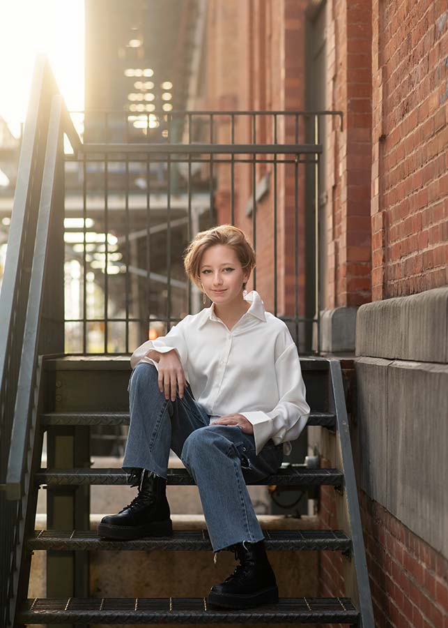 An Urban TriBeca high school senior with short hair, wearing a white shirt, blue jeans, and black boots, sits on a metal staircase outside a brick building, with soft sunlight in the background.