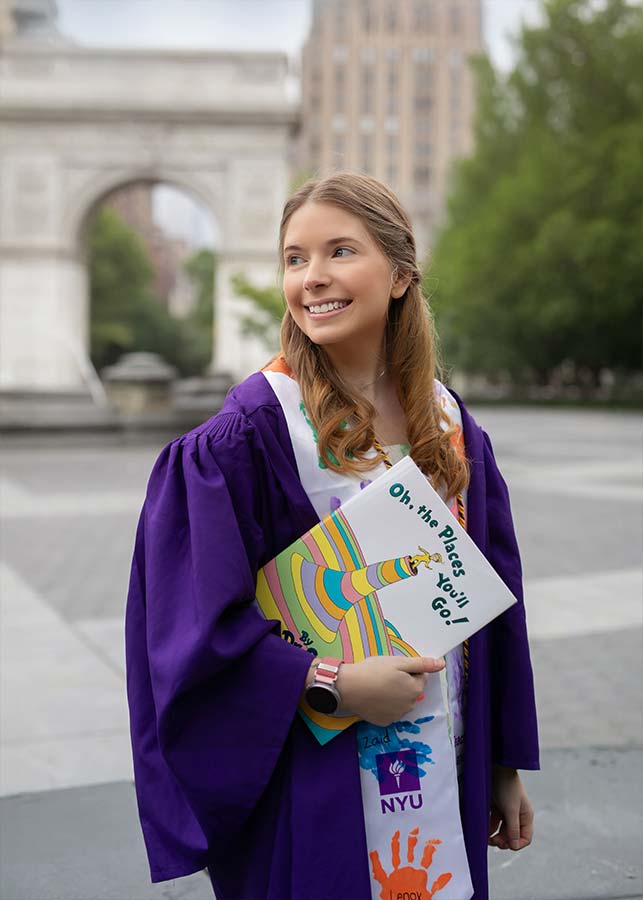 A young NYU graduate in a purple graduation gown stands outdoors, holding two colorful books, including