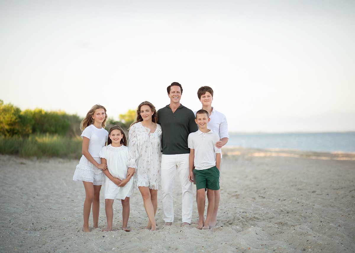 A family of six, dressed in white and green, stands barefoot on a sandy beach in Sag Harbor, NY, with lush greenery and the ocean in the background.