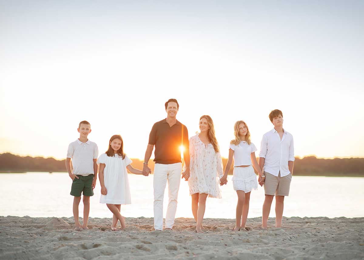 Six people, four children and two adults, stand on a sandy Sag Harbor, NY beach holding hands. The sun sets behind them over the water, casting a warm glow. They are all dressed in light-colored summer clothes.
