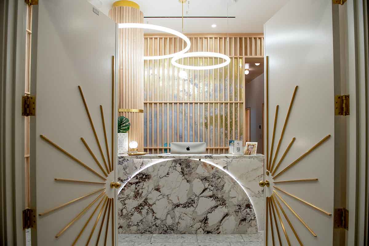A modern interior beauty salon reception area with a marble desk, backlit accents, and decorative wooden wall panels. Double doors with gold sunburst designs are open, revealing stylish circular ceiling lights above the desk.