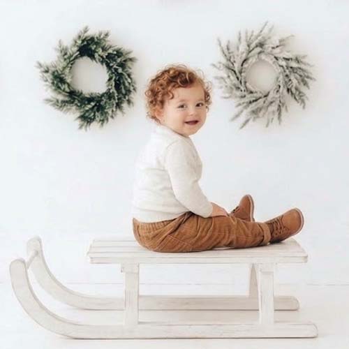 A toddler with curly hair sits on a white wooden sled, smiling, as if posing for a professional photographer. Two green wreaths hang on a white wall behind them.