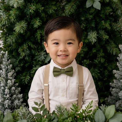 Young boy in a white shirt, tan suspenders, and green bow tie smiles while standing in front of lush green foliage and small frosted trees.