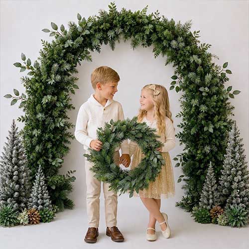 Two young children, a boy and a girl, stand smiling under a festive greenery arch with small frosted trees on either side. The girl holds a large green wreath. Both wear light-colored, dressy clothes.