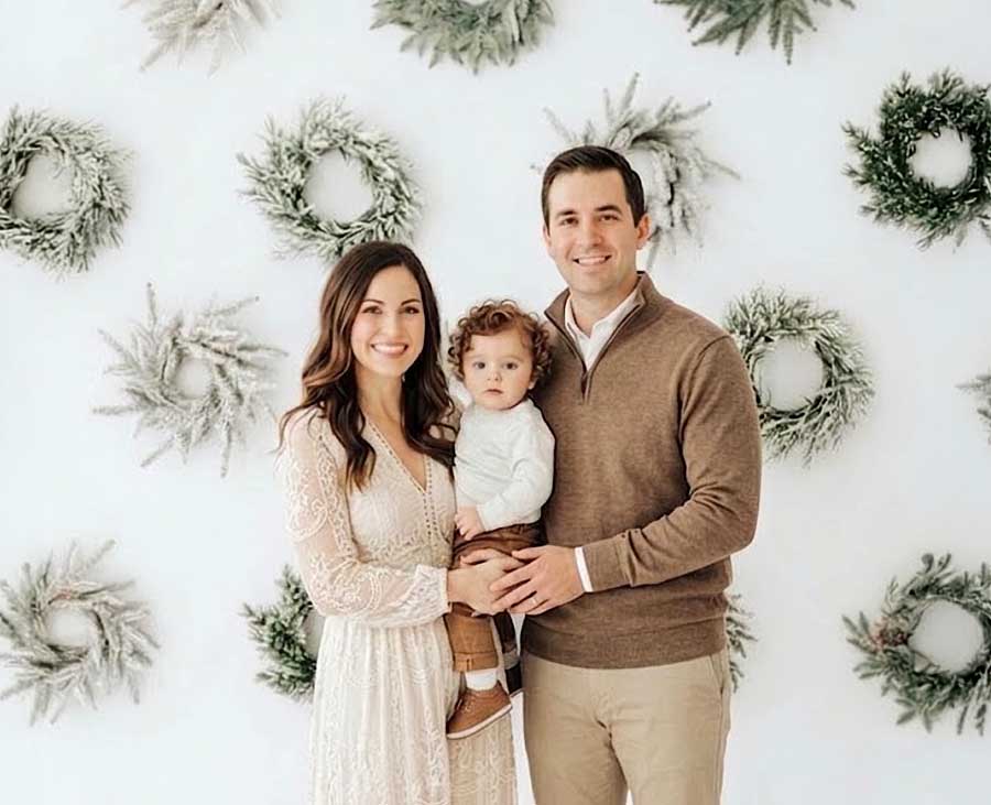 A smiling couple stands with their young child in front of a white wall decorated with frosted wreaths. The woman wears a white dress, the man a brown sweater, and the child a white sweater and brown boots.