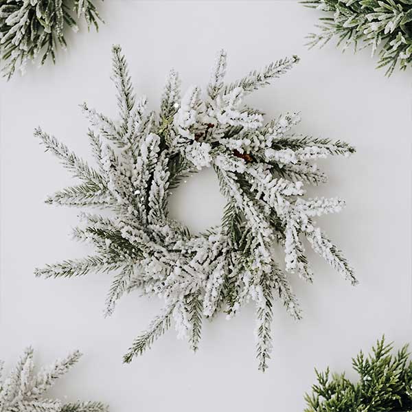 A frosted evergreen wreath with snow-like flocking is centered on a white background, surrounded by green, snow-dusted pine branches in each corner.
