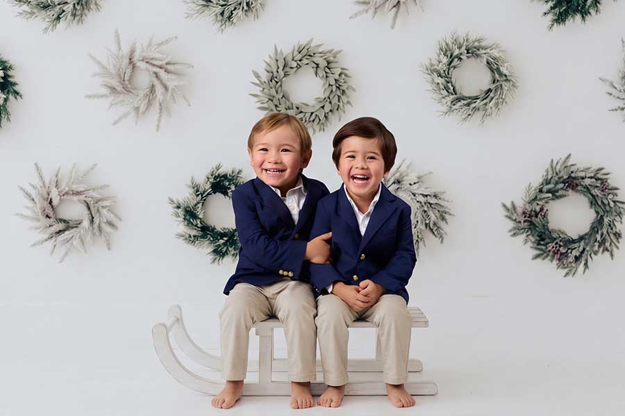 Two smiling young boys in navy blazers and khaki pants sit barefoot on a white sled, with frosted wreaths hanging on the white wall behind them.
