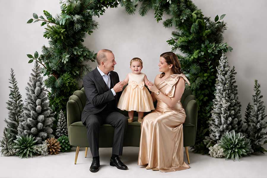 A family of three poses on a green couch, surrounded by greenery and frosted trees. The man wears a suit, the woman a gold dress, and the toddler in the middle wears a gold dress and headband, all smiling.